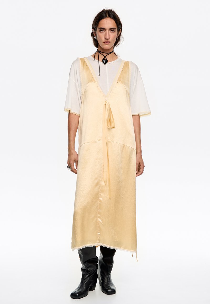 Abito giallo di satin con scollatura a V profonda e fiocco frontale, sovrapposto a una camicia bianca a maniche corte. Stivali neri al ginocchio completano il look.