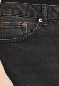 Los jeans de mezclilla negra presentan un diseño estructurado con herrajes de cobre y un bolsillo delantero visible. El material muestra una textura sutil.