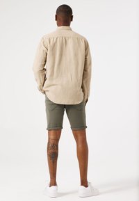 Beige linnen overhemd met lange mouwen, gecombineerd met olijfgroene shorts. Witte sneakers zichtbaar; tatoeage op de kuit heeft een bloemenontwerp.