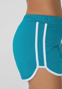 Shorts in cotone teal con bordo bianco. Presentano una vita aderente e un orlo curvo, evidenziando dettagli in piping laterali. Tessuto morbido.