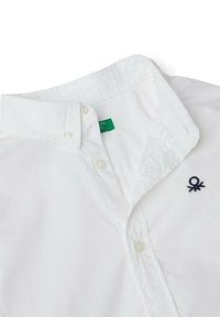 Camicetta bianca con bottoni, con un logo ricamato blu scuro sul petto e un'etichetta verde sul colletto. Texture liscia.