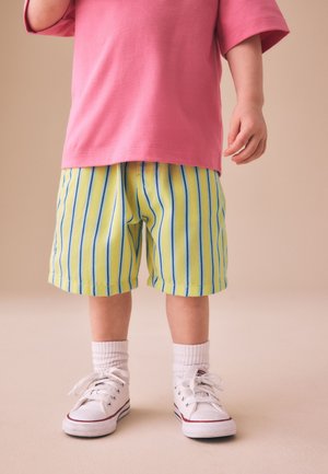 Kind trägt gelbe Shorts mit blauen vertikalen Streifen, weiße Socken, weiße Turnschuhe und ein pinkes kurzärmeliges Shirt und steht auf einem beigen Boden.