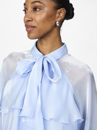Helles blaues Blusenshirt mit einer gebundenen Schleife am Kragen und rüschigen Lagen, aus einem glänzenden, halbtransparenten Material gefertigt. Dazu gehören silberne Tropfenohrringe.