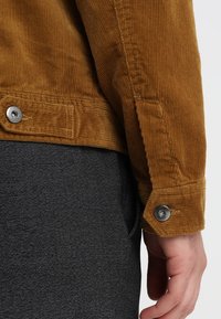 Brun kordfløjl jakke med en struktureret overflade, der har sølv trykknapper og en skræddersyet pasform. Bæres over mørke, strukturerede bukser.