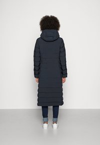 Marineblå lang pufferjakke med hætte, med en quiltet design, synlige sømme og lige pasform. Kombinationen med lyseblå jeans og hvide sneakers.