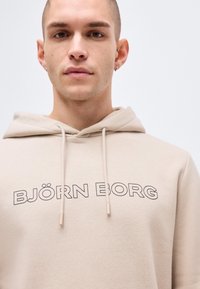 Νεαρός άνδρας με κοντά μαλλιά που φοράει μπεζ φούτερ με κουκούλα και την επιγραφή "BJÖRN BORG" στο στήθος σε απλό φόντο.