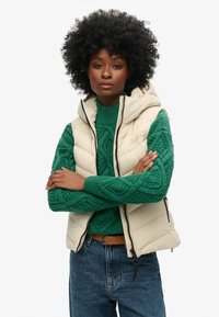 Gilet en duvet beige avec capuche, fermeture éclair à l'avant et détail de logo, porté sur un pull en maille torsadée vert, associé à un jean bleu et une ceinture marron.