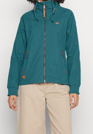 Tyrkysinsininen vetoketjullinen huppari huppulla ja kiristysnauhalla, vetoketjulliset etutaskut, beigejen housujen kanssa.