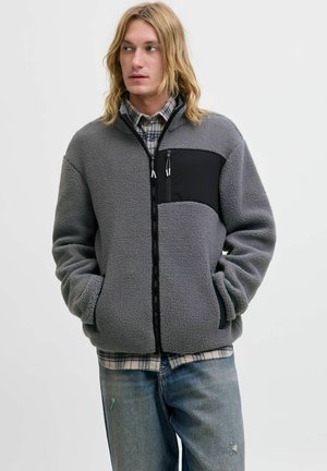 Grijze fleecjacket met zwarte zak, ritssluiting en zijzakken. Model draagt een geruit overhemd eronder en versleten jeans.