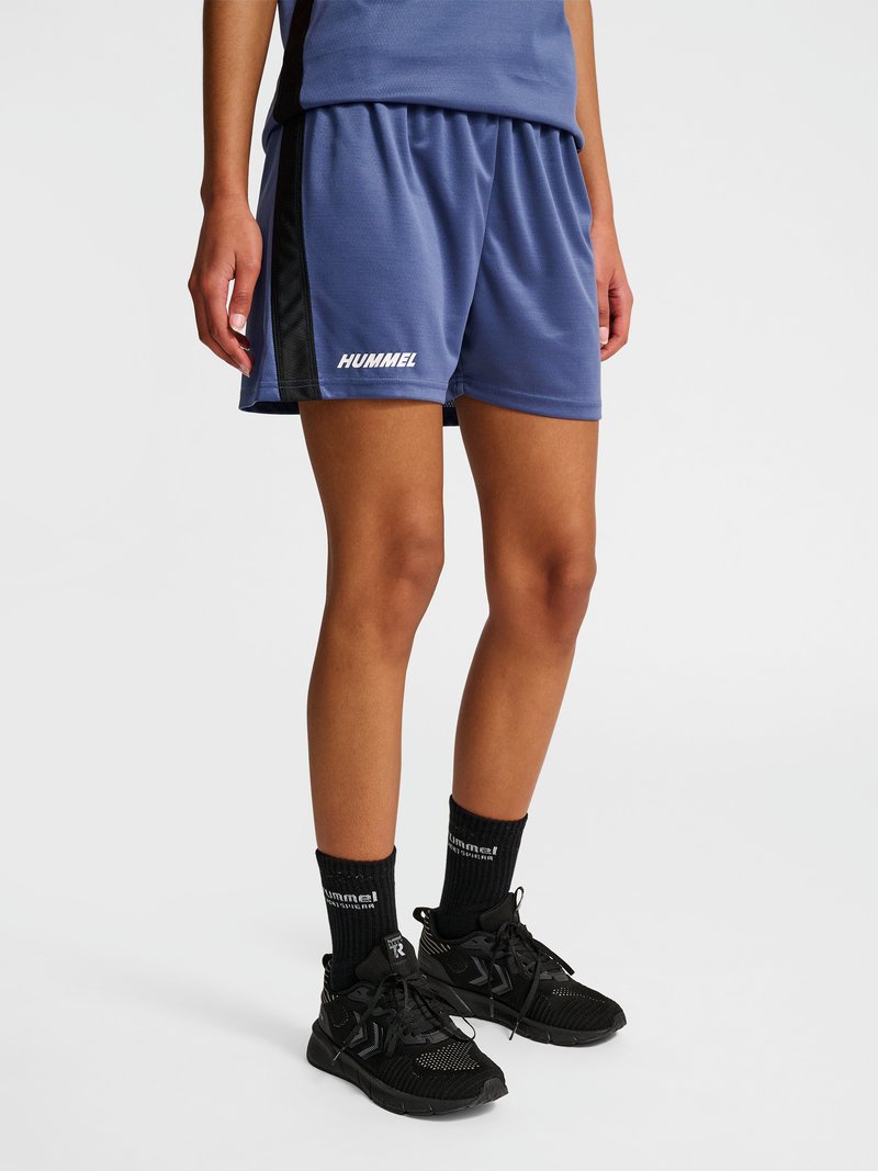 Personne portant un short de sport bleu avec des panneaux latéraux noirs, des chaussettes Hummel noires et des chaussures de sport noires, debout sur un fond blanc.