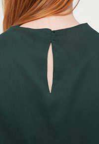 Tmavozelený top z hladkej tkaniny s kľúčovou dierkou na chrbte so zapínaním na gombík a jemne zaobleným výstrihom.
