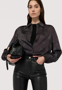 Czarna bluzka w wzory z jasnoniebieskimi konturami i zapięciem na guziki, zestawiona z wysokimi spodniami skórzanymi w kolorze czarnym, trzymająca czarną skórzaną torbę.