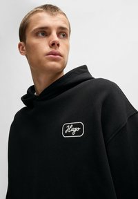 Felpa nera con cappuccio caratterizzata da un patch logo ricamato in bianco sul davanti. Il tessuto ha una texture liscia, con un design rilassato e una vestibilità oversize.
