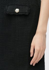 Gros plan sur une main reposant à côté d'une jupe en tissu texturé noir avec un rabat de poche orné d'un grand bouton en perle.