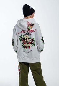 Sudadera blanca con un gráfico de calavera y rosa en la parte posterior, con acentos en verde y rojo. Mangas largas decoradas con diseños similares.