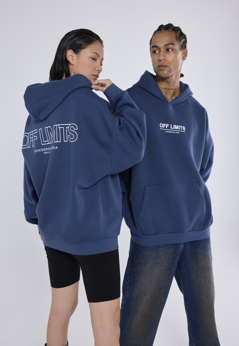 Sudaderas de color azul marino con texto blanco que dice "ÁREA PELIGROSA PROHIBIDA" en la espalda y el frente, con un bolsillo canguro y un corte relajado.