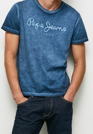 Homme portant un t-shirt bleu Pepe Jeans London et un jean en denim foncé, les mains dans les poches avant.