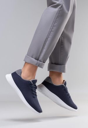 Marineblauwe gebreide sneakers met grijze vilten accenten, witte zolen en vetersluiting, zwevend in de lucht met opgerolde grijze broek.