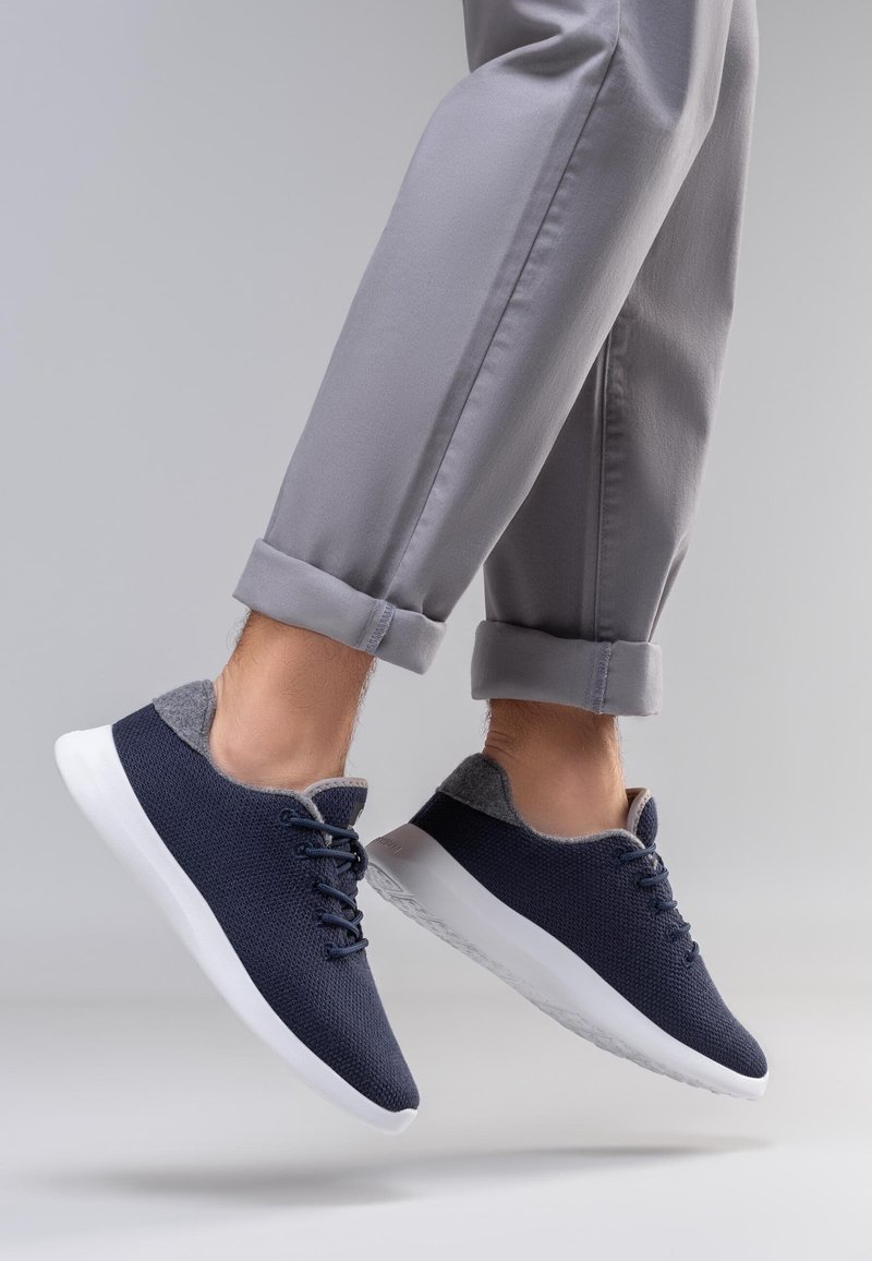 Baskets en maille bleu marine avec accents en feutre gris, semelles blanches et design à lacets, présentées en l'air avec un pantalon gris retroussé.