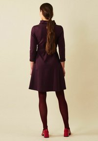 Burgundin ruudullinen mekko, jossa on kaulus, kolmemetriset hihat ja vyö vyötäröllä; yhdistettynä marooninvärisiin sukkahousuihin ja punaisiin korkokenkiin.