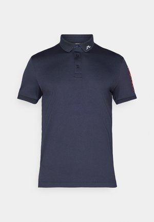 Polo azul-marinho de mangas curtas, com botão de três lugares e logótipo no colarinho. Apresenta texto em destaque vermelho na manga esquerda.