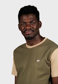 Camiseta de manga corta en verde oliva y beige con cuello redondo y un pequeño logo de elefante bordado en el pecho. Tejido de algodón suave.