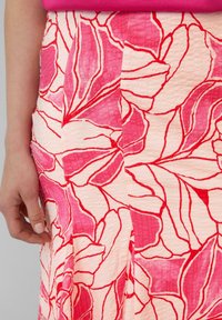 Jupe florale en tissu léger rose pâle texturé, ornée de motifs de feuilles rouges et roses audacieux. Le design présente des lignes épurées et une légère brillance.