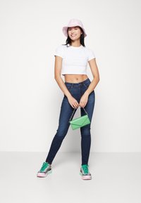 Mlada žena u bijelom skraćenom majici, tamnim uskim trapericama, ružičastom šeširu s krovom i šarenim tenisicama, drži malu zelenu torbicu.