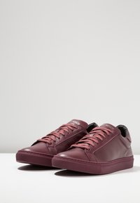 Burgundy leather sneakers with flat laces, a rounded toe, rubber outsole, and stitched detailing. Branding on tongue. Smooth texture.