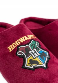 Pantofole in velluto borgogna con ricamo del crest di Hogwarts, dettagli blu, verdi, rossi e gialli, e la scritta "HOGWARTS" in oro.