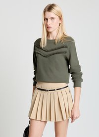 Sweat-shirt court vert olive avec lignes chevrons texturées et manches longues, associé à une mini-jupe plissée beige et une ceinture noire.