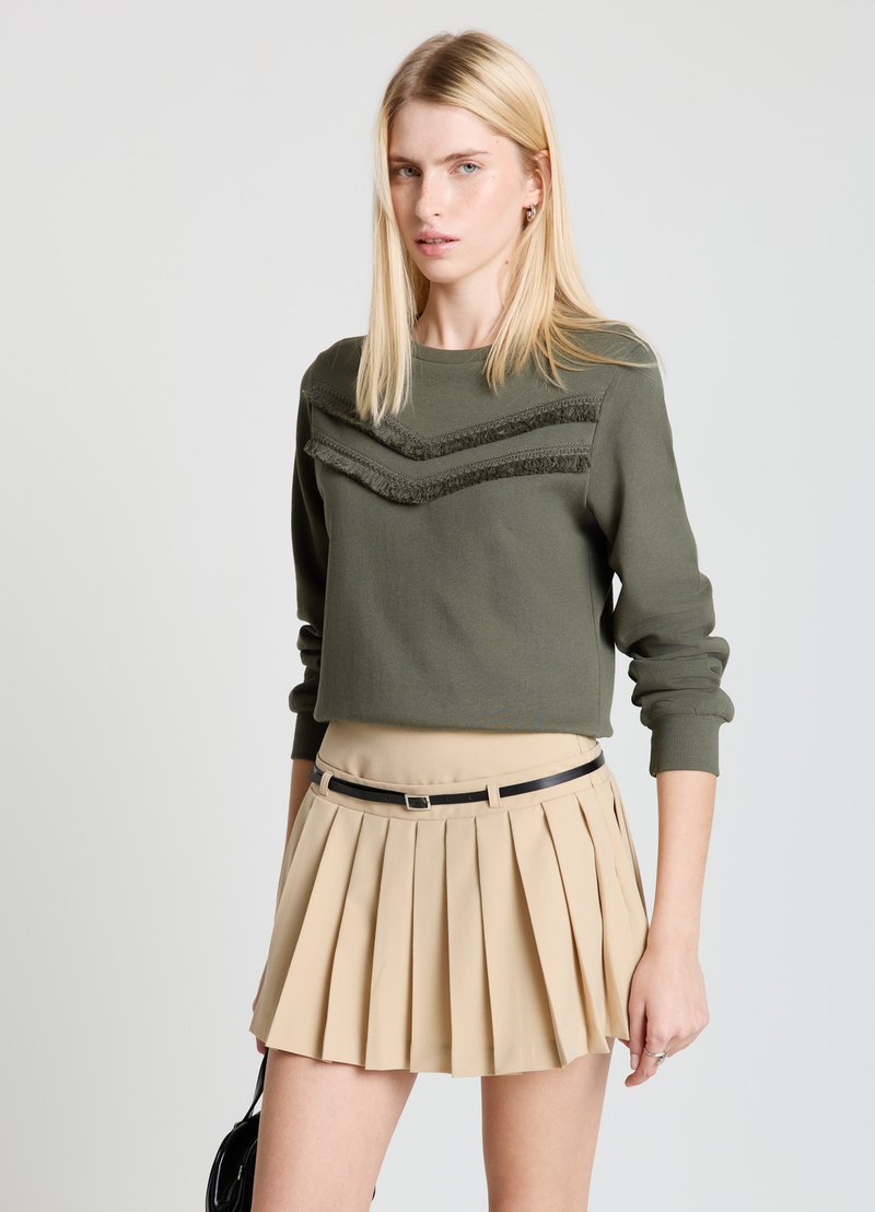 Sweat-shirt court vert olive avec lignes chevrons texturées et manches longues, associé à une mini-jupe plissée beige et une ceinture noire.