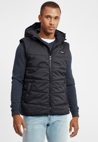 Schwarze gepolsterte Weste mit Kapuze, Reißverschluss vorne und zwei Seitentaschen. Getragen über einem dunkelblauen Langarmhemd und hellen Jeans.