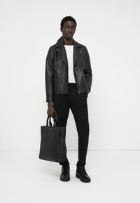 Svart skinnbikerjakke over en hvit skjorte, parret med smale svarte bukser og svarte boots. Holder en svart tote-bag med vertikale aksenter.