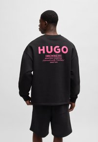 Felpa nera con stampa rosa bold sulla schiena con scritto "HUGO" e altri dettagli. Design casual, maniche lunghe e orlo a costine.