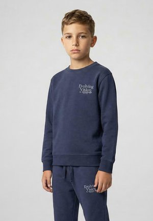Jongen staat en draagt een bijpassende marineblauwe trui en joggingbroek met de tekst "Evolving Vision" op beide kledingstukken.
