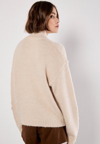 Crèmekleurige, chunky gebreide trui met een hoge hals, verlaagde schouders en ribbelzoom. Zachte textuur en een relaxte pasvorm.