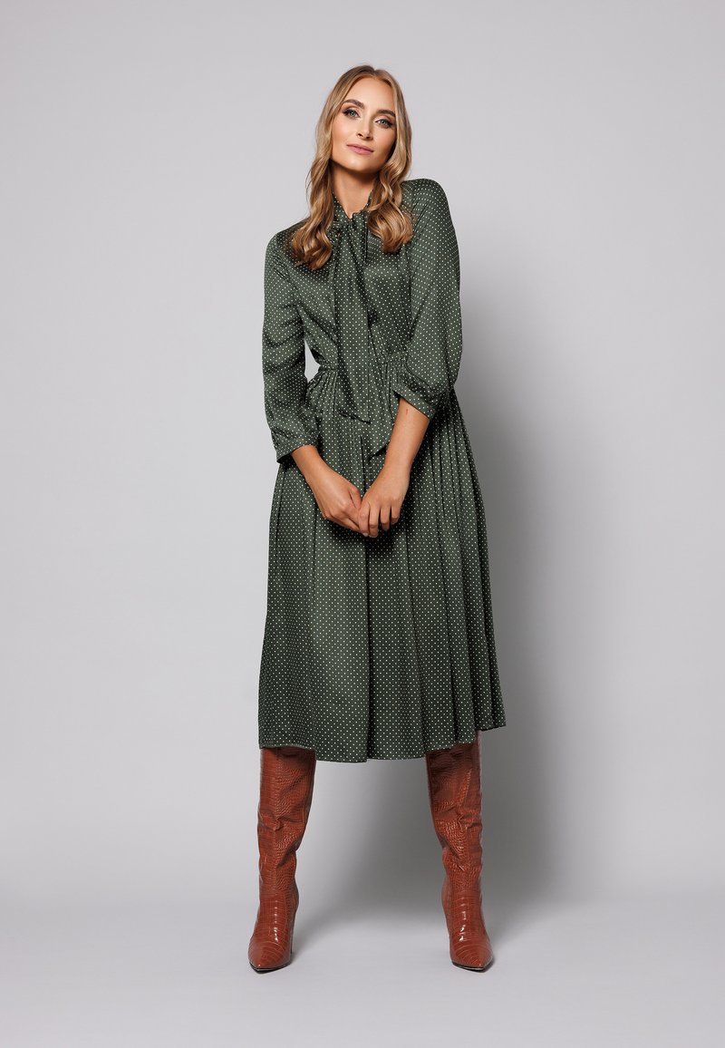 Zielona sukienka w kropki z wiązanym dekoltem, trzyczwartowymi rękawami i rozkloszowanym dołem. Dobrana z błyszczącymi, brązowymi kozakami za kolano.