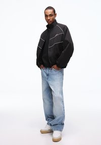 Black bomber jacket with white stitching, worn over a black sweater, paired with light blue baggy jeans and white sneakers with beige soles.