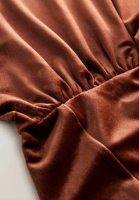 Brown velvet fabric with a soft, smooth texture. Gathered sections create folds, highlighting draping and fluidity in the material.