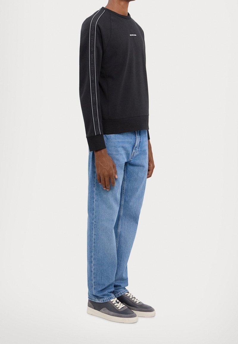 Homme portant un sweat-shirt Michael Kors noir à manches longues avec une bande logo sur les manches, un jean bleu clair et des baskets grises à lacets, debout de profil.