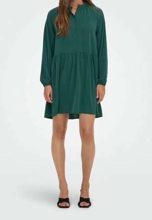 Femme portant une robe ample à manches longues vert foncé avec des boutons et des sandales à talons noires à bout ouvert, debout sur fond blanc.