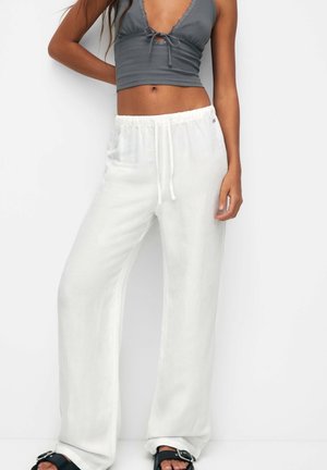 Vrouw draagt losse witte broek met trekkoord, grijs mouwloos cropped topje met knoopdetail aan de voorkant en zwarte sandalen met gesp.