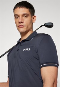 Ναυτικό πόλο μπλουζάκι από λεία ύλη, με λευκό λογότυπο, πλέξιμο με δύο κουμπιά και αντίθετες ρίγες στους ώμους. Γκολφ κλαμπ στο βάθος.