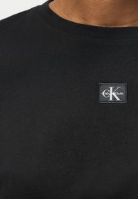 Κοντινό πλάνο μαύρου t-shirt με στρογγυλή λαιμόκοψη και μικρό τετράγωνο λογότυπο Calvin Klein στο αριστερό στήθος.