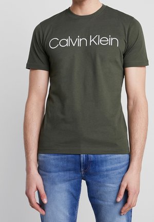 Mann trägt ein olivgrünes kurzärmeliges Hemd mit weißem Calvin Klein Logo, kombiniert mit blauen Jeans, vor einfachem grauem Hintergrund.