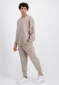 Felpa beige chiaro e pantaloni jogger abbinati realizzati in tessuto morbido; presenta una tasca con zip e una patch con il logo; abbinati a sneaker bianche.