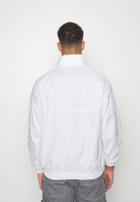 Vit vindjacka med hög krage, elastiska cuffs och fåll. Slät textur med subtil sömdetalj på baksidan.