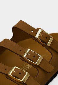 Καφέ λουριά σανδαλιών με ρυθμιζόμενες αγκράφες σε χρυσό τόνο που εμφανίζουν χάραξη της μάρκας, κοντινό πλάνο σε μαλακό υλικό με υφή και οπές για το κλείσιμο.