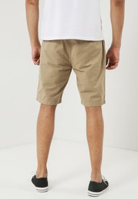 Shorts en coton beige avec une coupe régulière, longueur genou, dotés d'une poche arrière et d'un ourlet replié, associés à des chaussures en toile noires.
